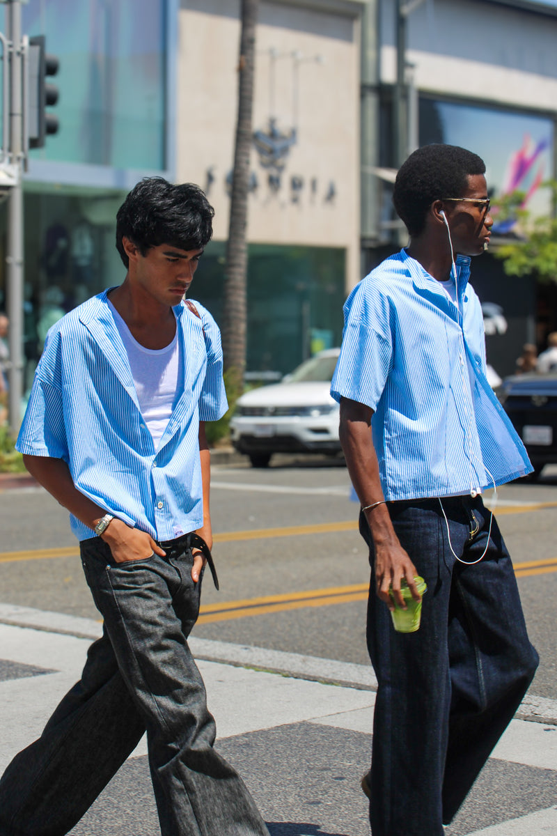 Two men in blue shirts crossing a street with cars and buildings in the background.