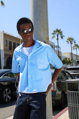 Man wearing a light blue shirt and sunglasses on a city street with palm trees in the background.
