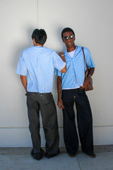 Two men in matching light blue shirts and dark pants standing against a white wall.