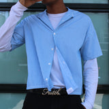 Person wearing a blue checkered shirt over a white long-sleeve shirt, standing in front of a glass door.
