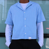 Person wearing a blue and white striped shirt over a white long-sleeve shirt, standing in front of a glass door.