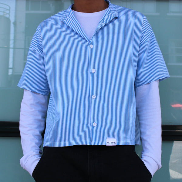 Person wearing a blue and white striped shirt over a white long-sleeve shirt, standing in front of a glass door.